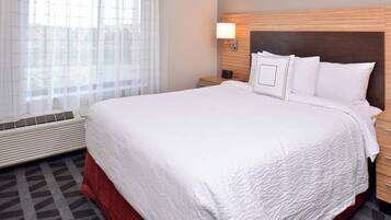 A hotel room with a large bed, a bedside table, a nightstand, a wall-mounted lamp, and a window with sheer curtains.