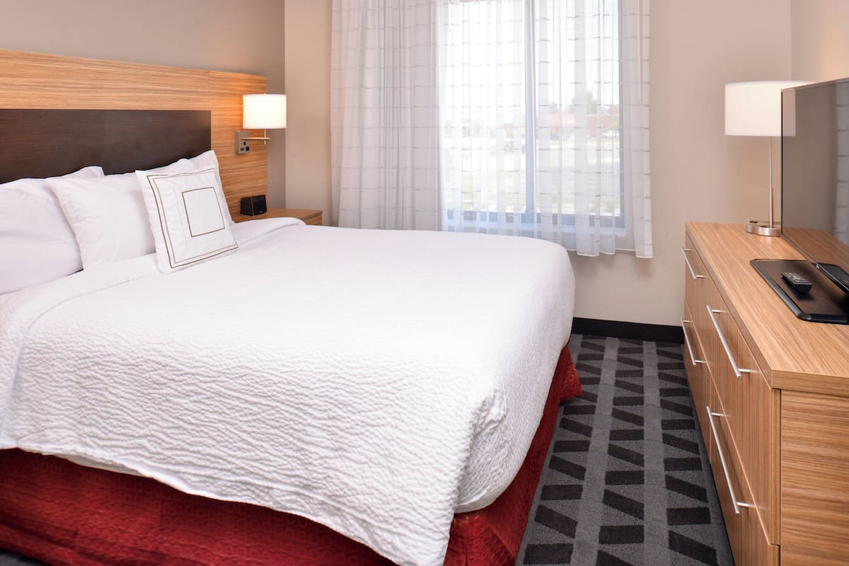 A hotel room with a large bed, a wooden headboard, a bedside table with a lamp, a TV, and a patterned rug.