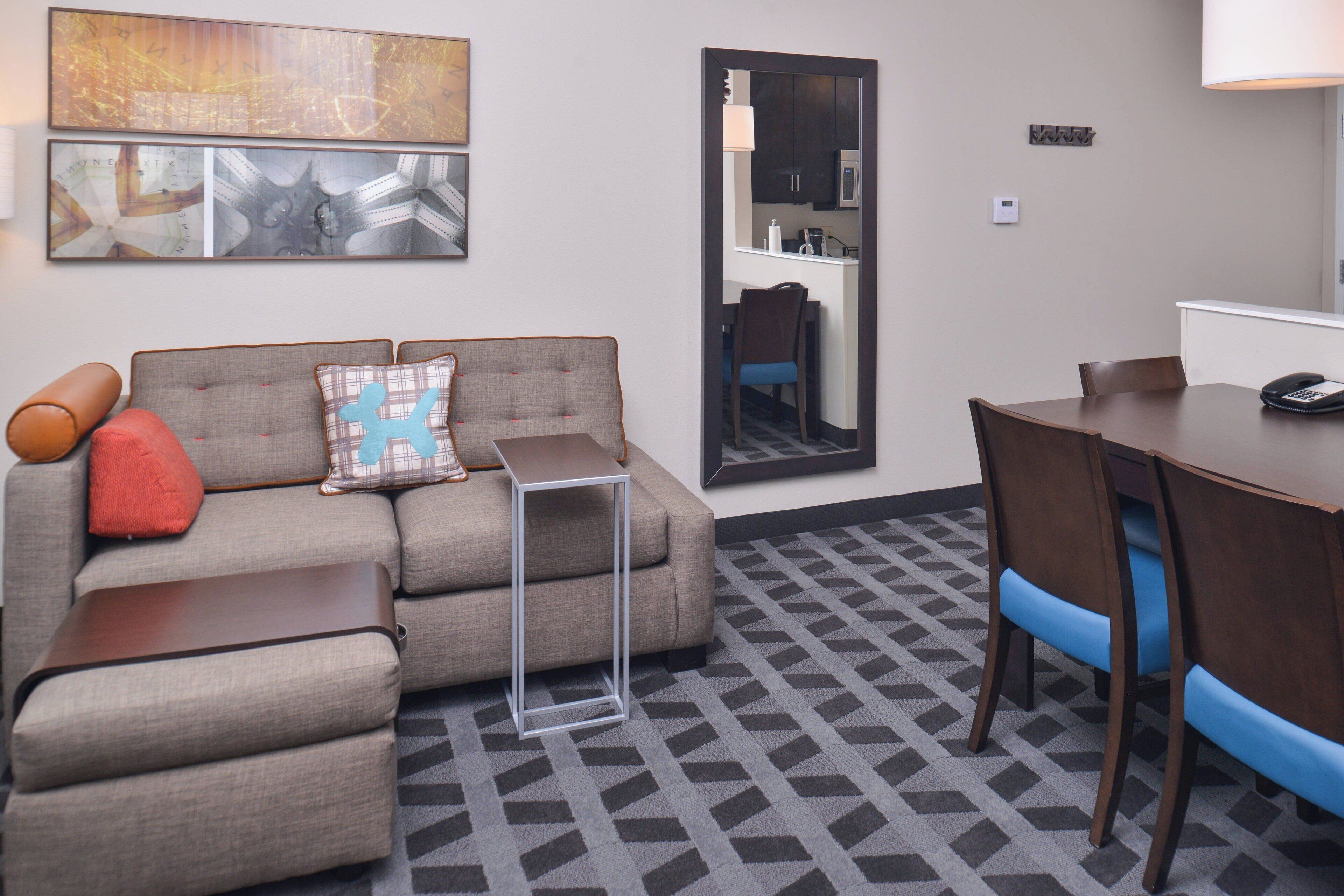 Ein Hotelzimmer mit einer Couch, einem kleinen Tisch, einem Esstisch mit Stühlen, einem Spiegel und einem durch den Spiegel sichtbaren Küchenbereich.