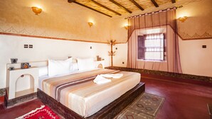 A bedroom with a large bed, a wooden headboard, a small bedside table, a nightstand, a decorative wall hanging, a window with curtains, and a patterned rug. - Ecolodge l Ile de Ouarzazate (Tarmigt)