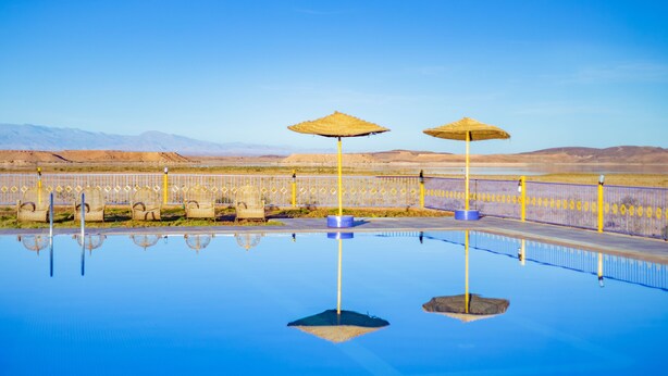 A pool with two umbrellas, a fence, and a desert landscape in the background. - Ecolodge l Ile de Ouarzazate (Tarmigt)