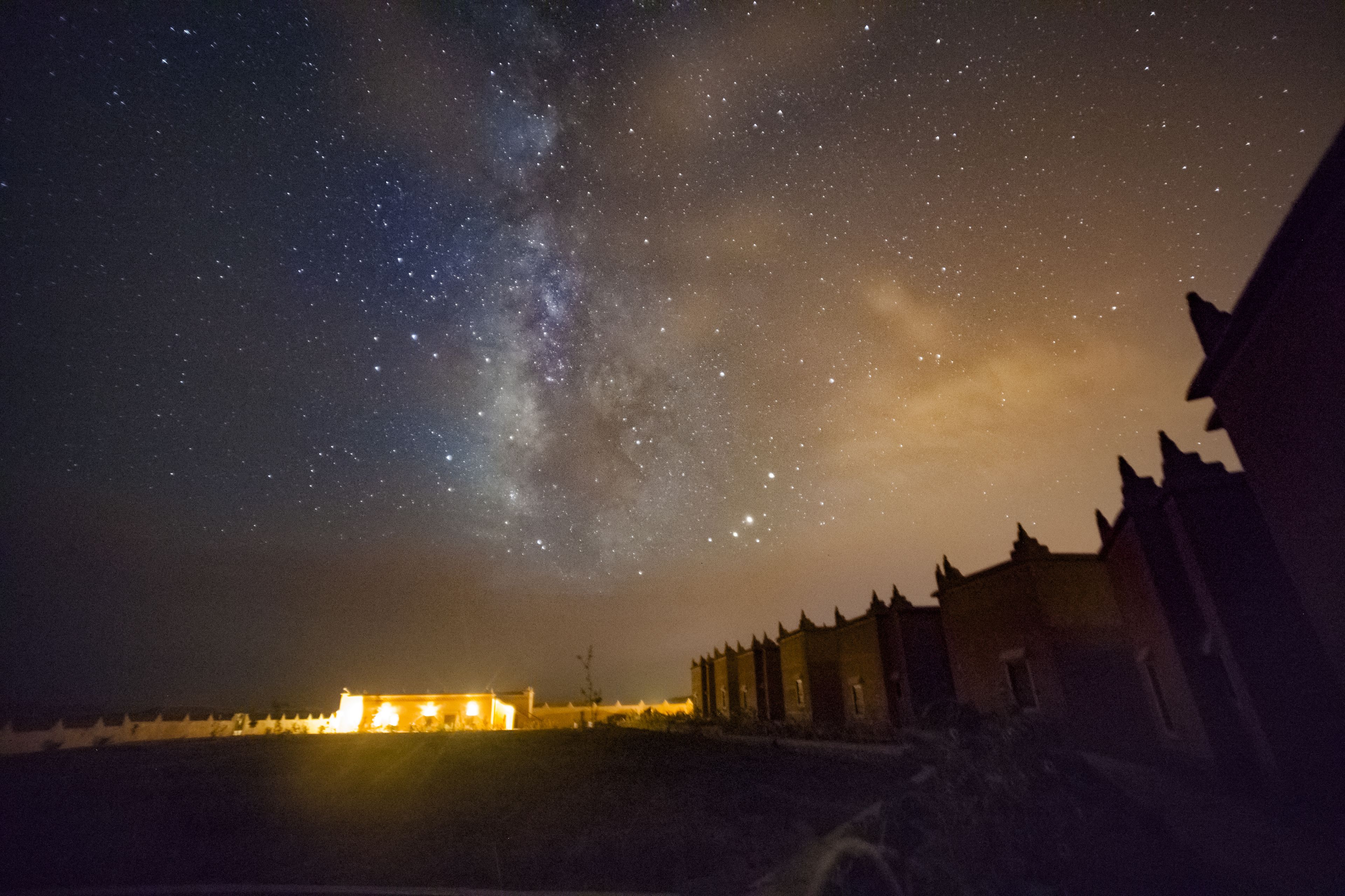 A night sky with stars and the Milky Way over a historic building with a lit interior.