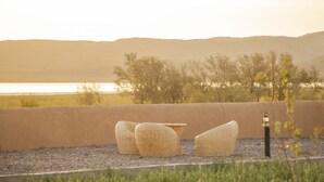 A seating area with wicker chairs and a table, overlooking a body of water and hills. - Ecolodge l Ile de Ouarzazate (Tarmigt)