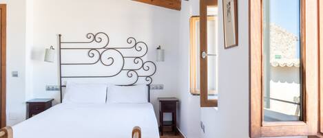 A bedroom with a wooden headboard, a bed with white linens, two bedside tables, a nightstand, a window with a view, and a wall-mounted light fixture.