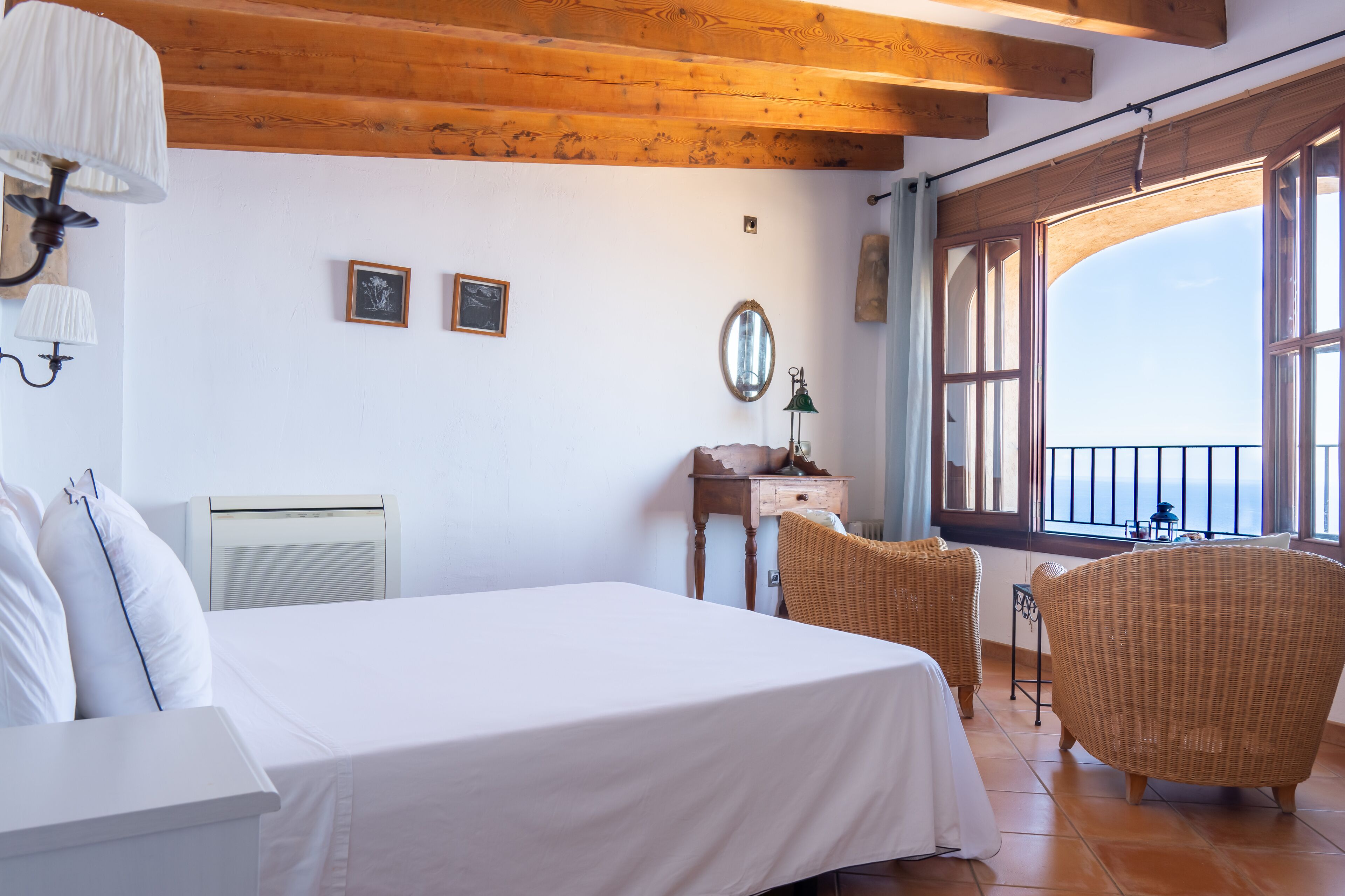 A bedroom with a bed, two wicker chairs, a dresser, a mirror, and a window with a view.