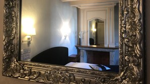 A mirror with an ornate frame reflecting a room with a bed, a nightstand, and a wall-mounted lamp. - Hôtel Les Deux Magots (La Roche-Bernard)