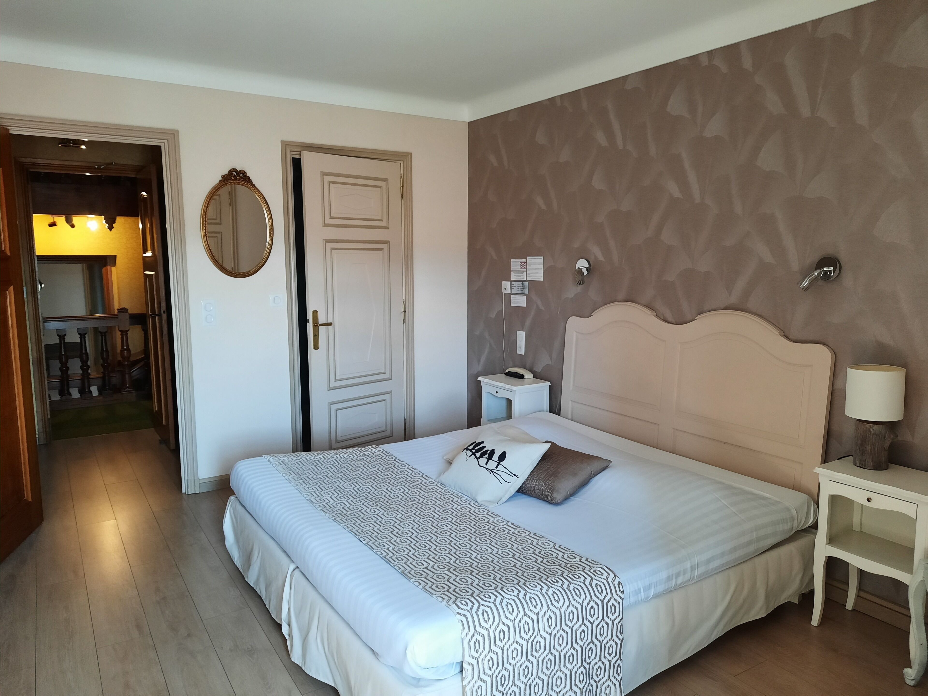 Una camera d'albergo con un letto grande, un comodino con una lampada, uno specchio a parete e una porta che dà su un corridoio.