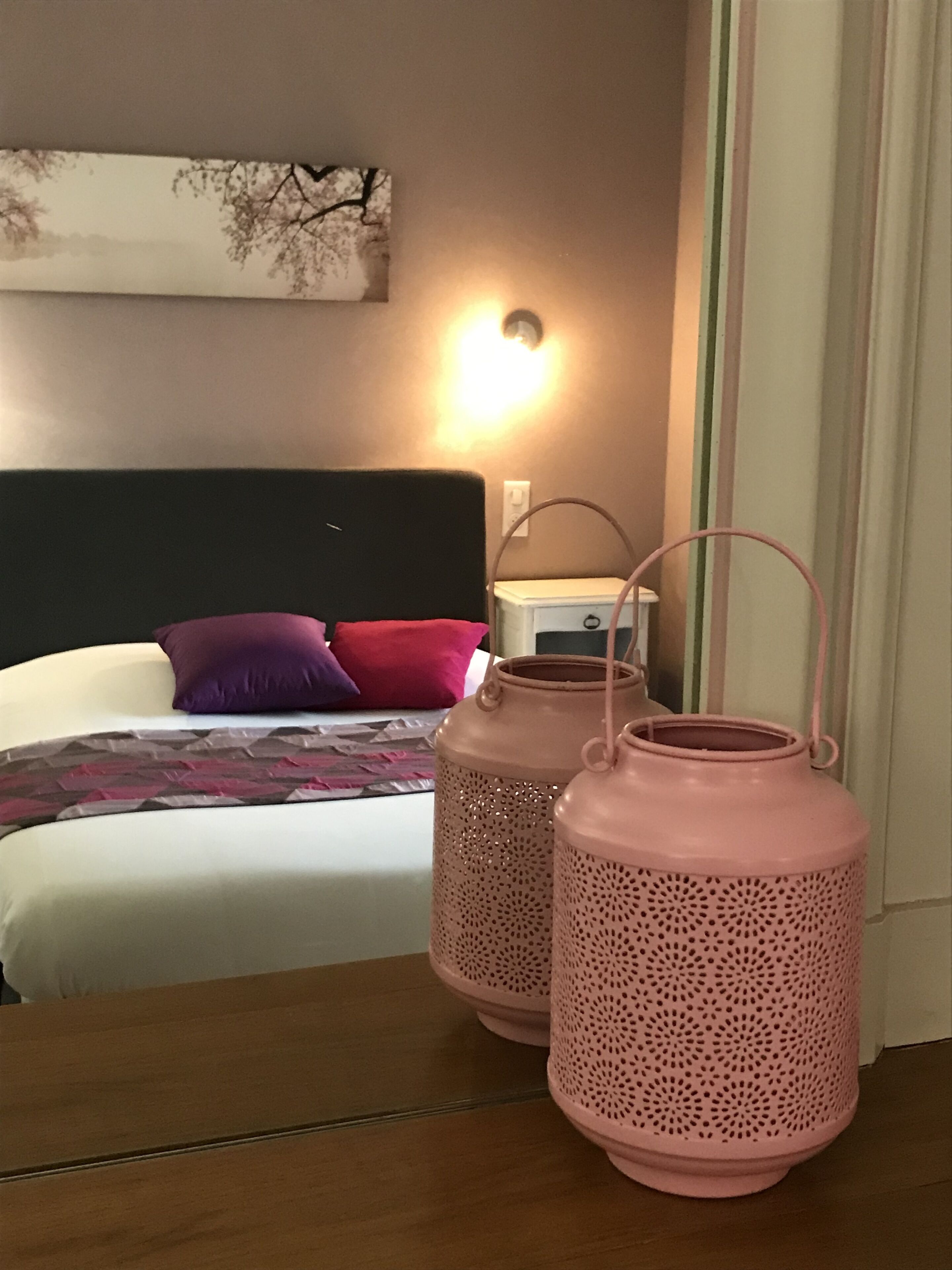 Une chambre à coucher avec un lit, des oreillers, une table de chevet et deux lanternes roses posées sur une table en bois.