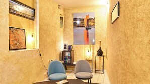 A narrow hallway with a tiled floor, two blue chairs, a small table, and a decorative rug. - Hôtel Les Deux Magots (La Roche-Bernard)