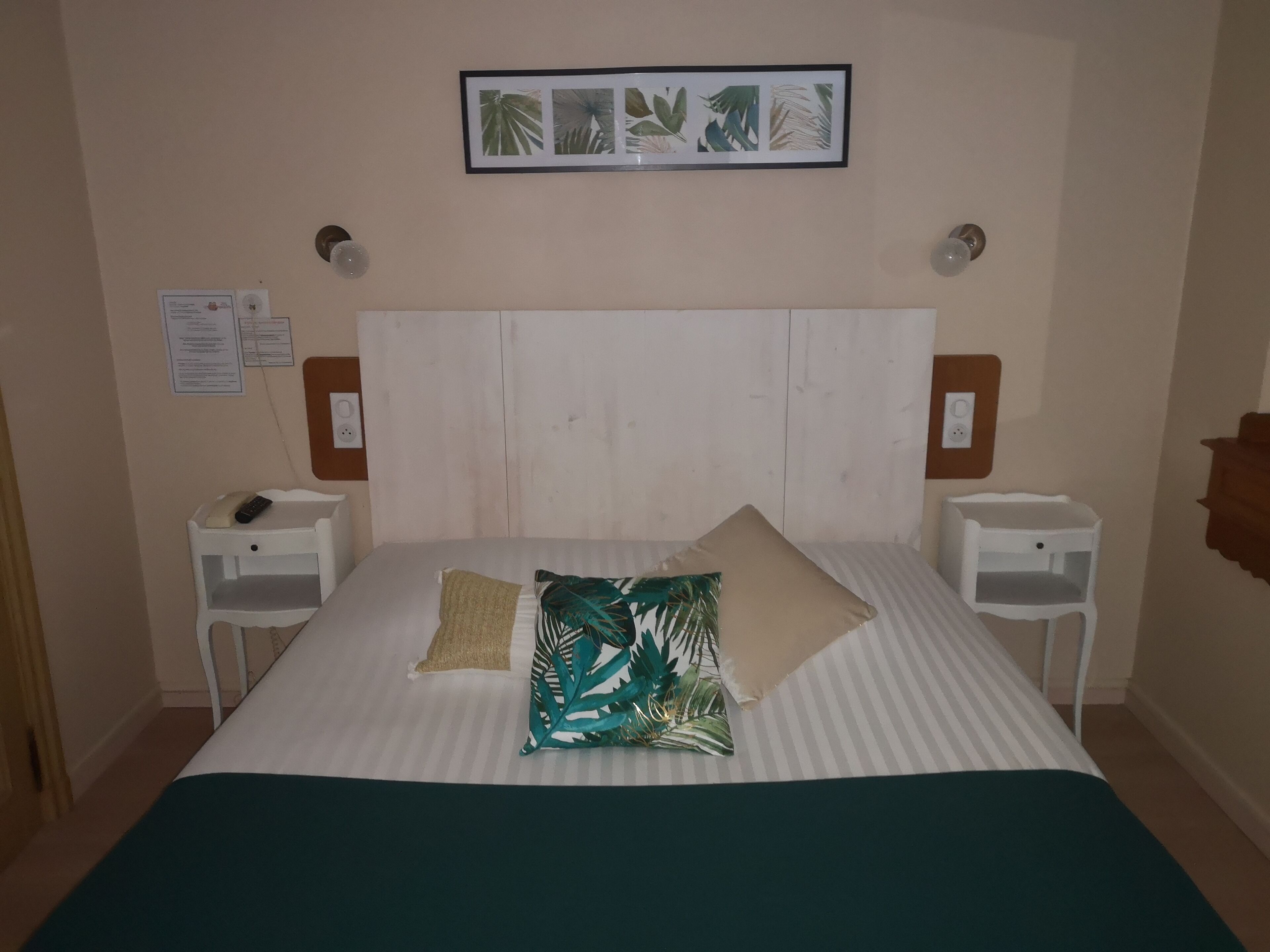 Un lit bien fait, recouvert d’une couvre-lit à rayures et agrémenté de deux oreillers, dont l’un est orné d’un motif de feuilles tropicales. Deux tables de chevet avec des lampes se trouvent de part et d’autre du lit, et une estampe botanique encadrée est accrochée au-dessus.