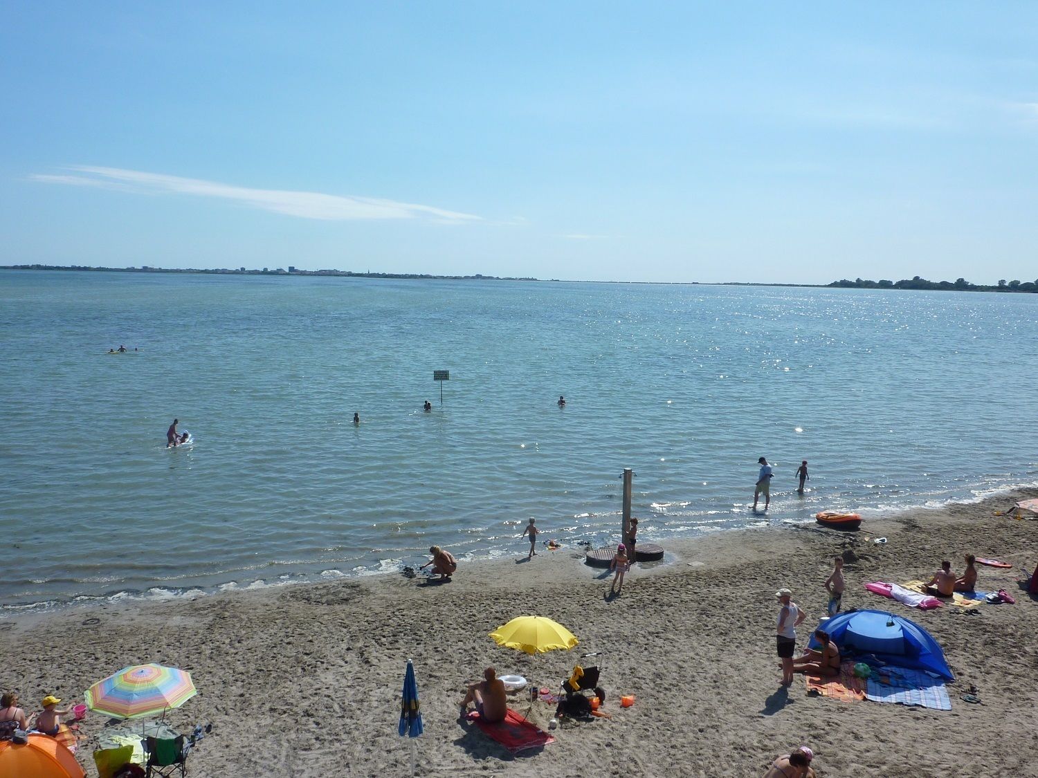 Ein Strand mit badenden und sonnenden Personen. Sonnenschirme und Liegen stehen im Sand.