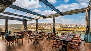 Een terras met tafels en stoelen, grote ramen bieden uitzicht op een stadsgezicht en een rivier.