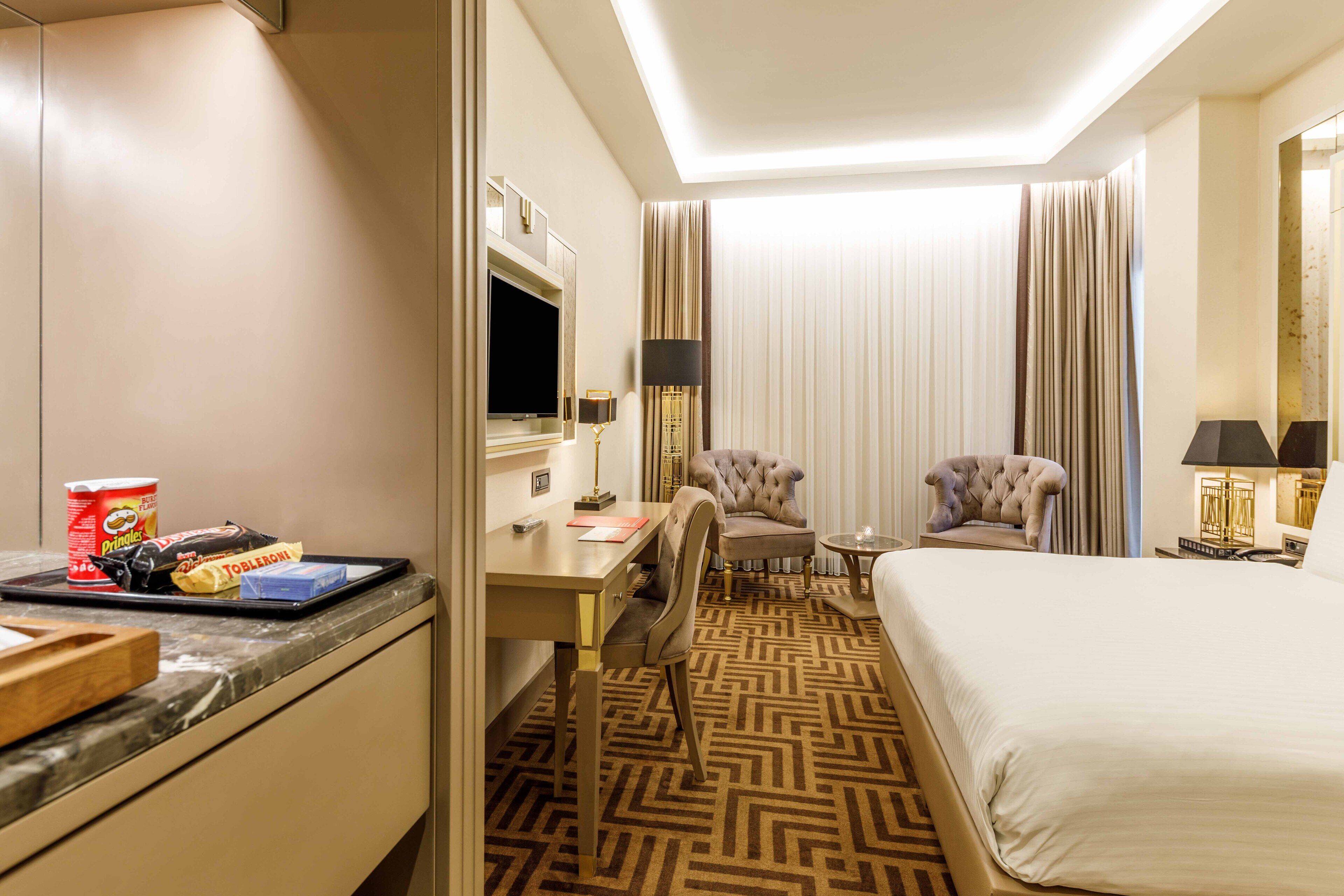A hotel room with a bed, two armchairs, a desk with a television, and a minibar.