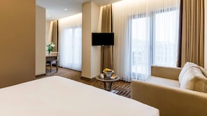Een hotelkamer met een groot bed, een sofa, een televisie, een klein tafeltje met fruit en een uitzicht op de stad door grote ramen.
