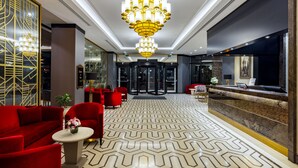 Een hotel lobby met een grote kroonluchter, rode zitplaatsen, een receptiebalie en een vloer met patroon.