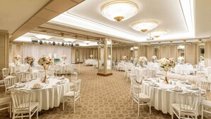 Een feestzaal met ronde tafels gedekt voor evenementen, met kroonluchters en bloemstukken als decoratie.