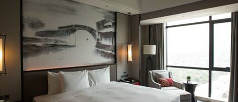 A hotel room with a large bed, a chair, a desk, and a large window offering a view.