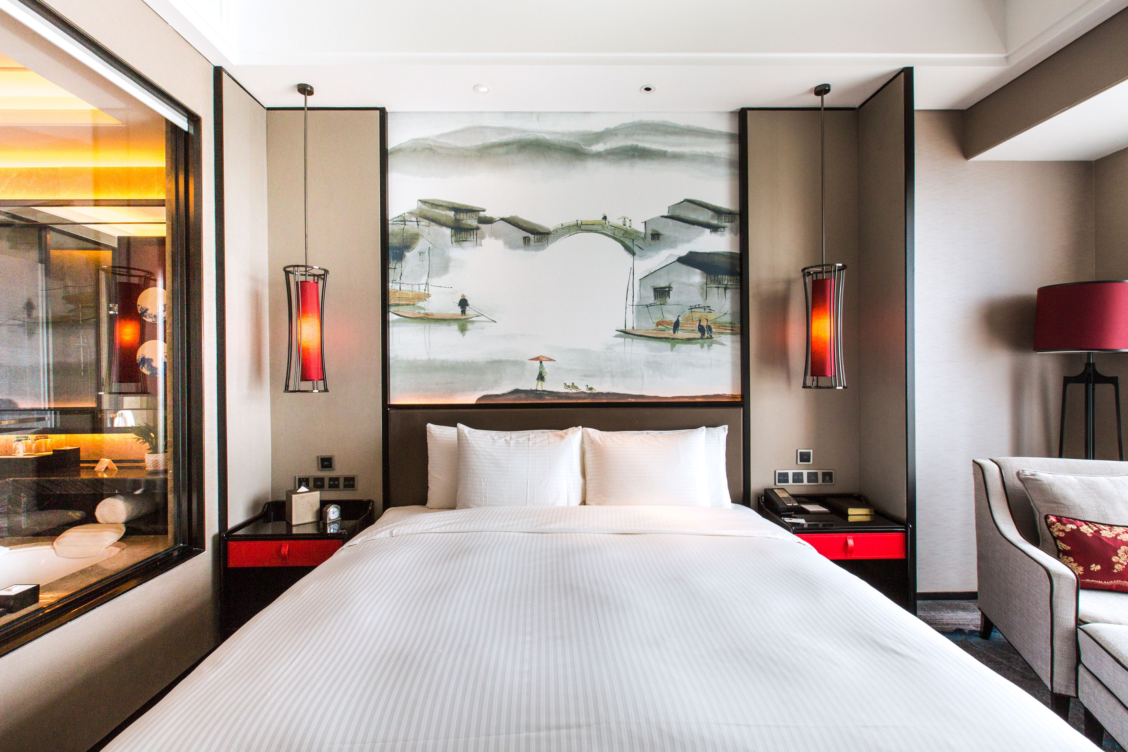 Habitación de hotel con una cama grande, una mesita de noche con una lámpara, un cuadro de un puente y vistas de un paisaje urbano a través de una ventana.