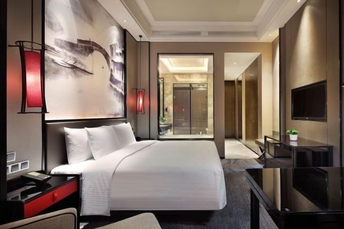 A modern hotel room with a large bed, a desk with a computer, a flat-screen TV, and a glass door leading to a balcony.