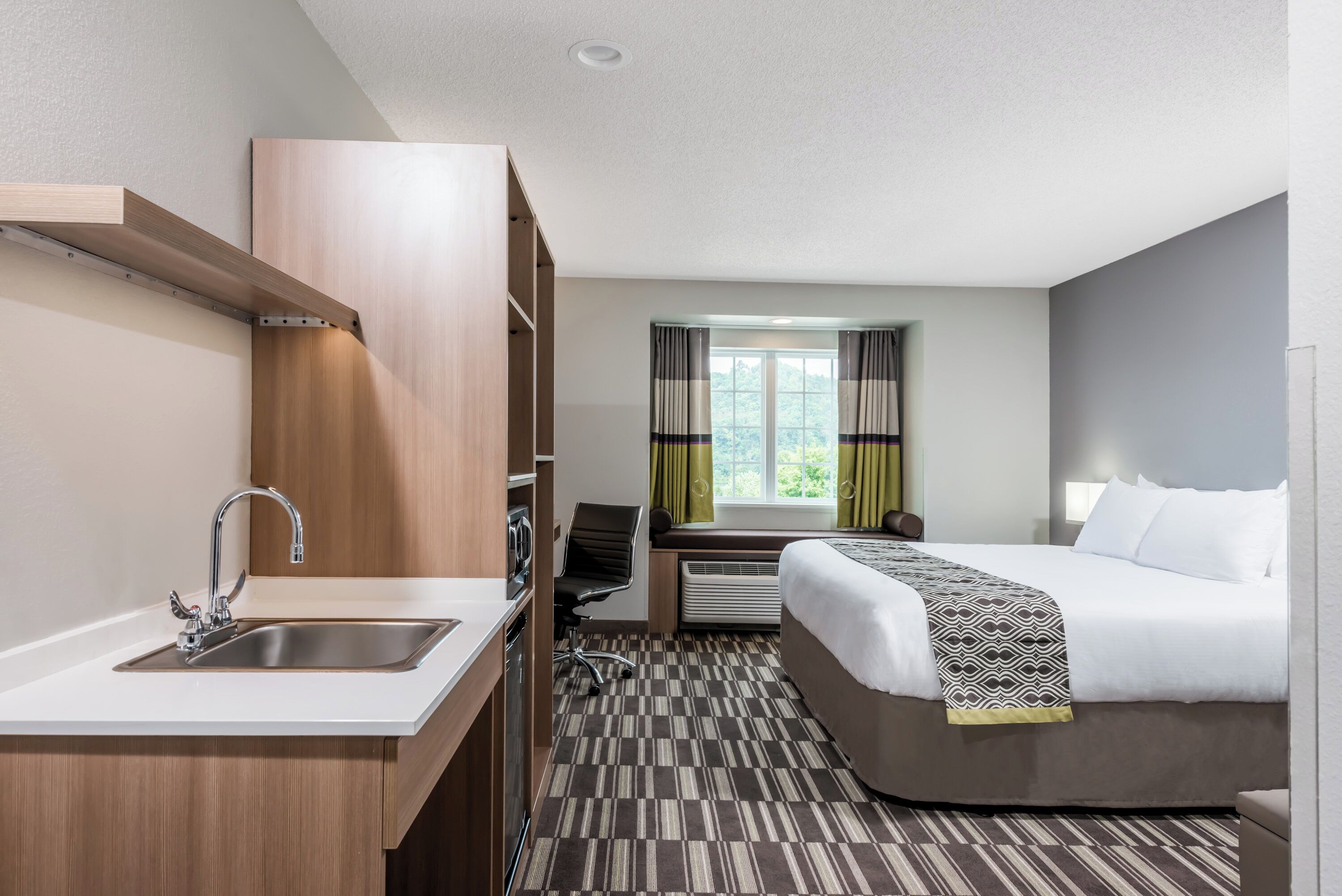 A hotel room with a kitchenette, a bed, a window with curtains, and a carpeted floor.