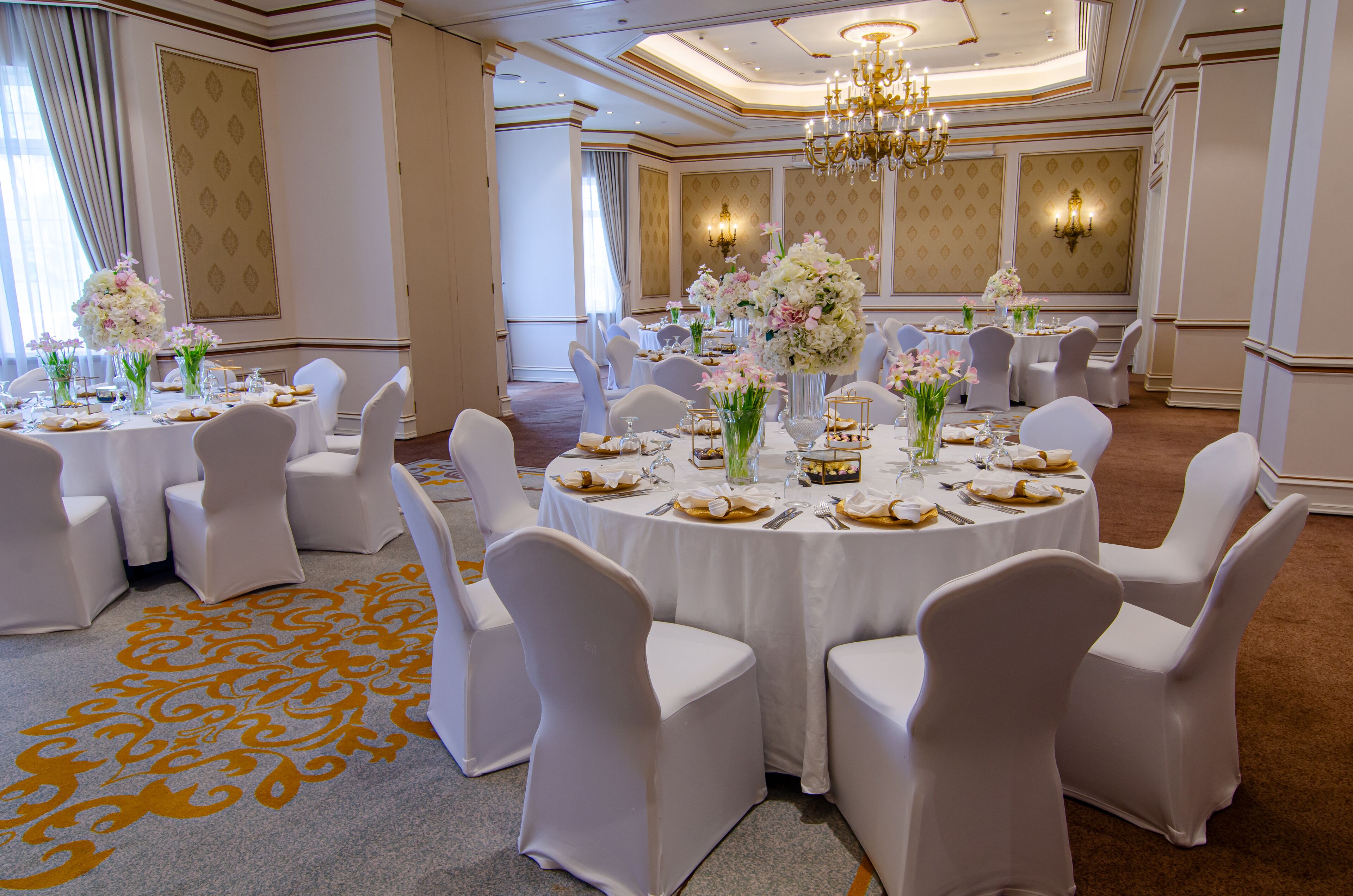 Un salón de banquetes con mesas redondas, sillas blancas y centros de mesa florales.