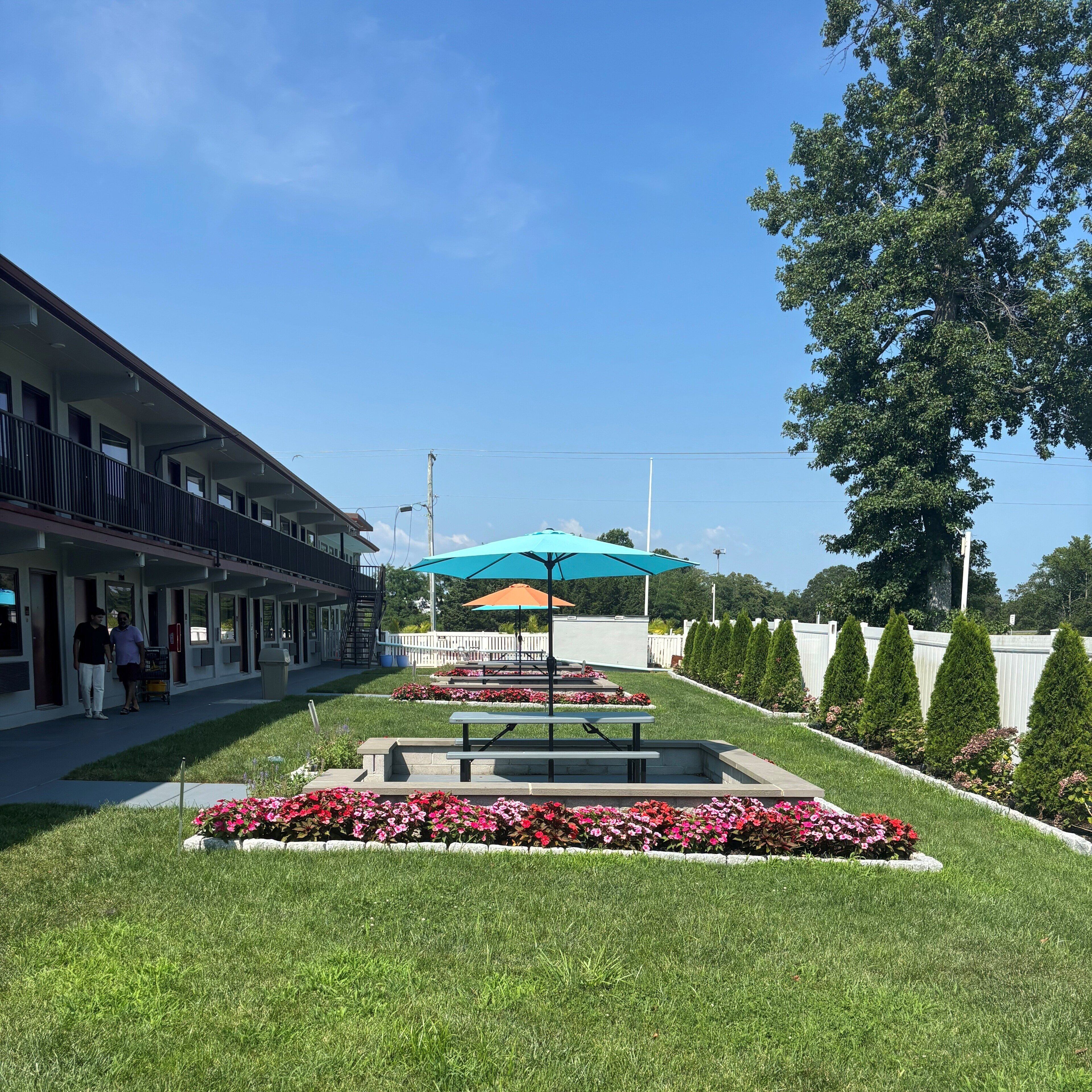A hotel with a covered outdoor seating area, a well-maintained lawn, and a row of neatly trimmed trees.