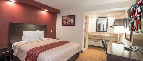 A hotel room with a bed, a desk with a chair, a mirror, and a bathroom visible through an open door.