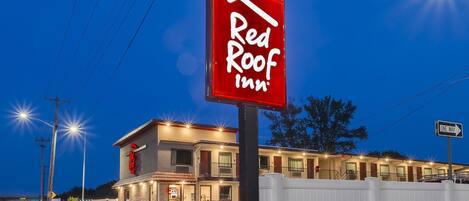Un hôtel Red Roof Inn reconnaissable à son grand panneau rouge, à son bâtiment bien éclairé et à son parking délimité par une clôture.