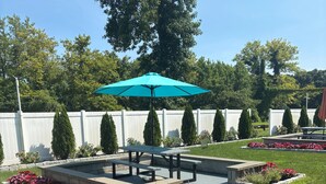 A patio area with a picnic table and a turquoise umbrella, surrounded by flower beds and a white fence. - Red Roof Inn Wildwood - Cape May/ Rio Grande (Rio Grande)
