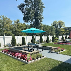 A patio area with a picnic table and a turquoise umbrella, surrounded by flower beds and a white fence. - Red Roof Inn Wildwood - Cape May/ Rio Grande (Rio Grande)