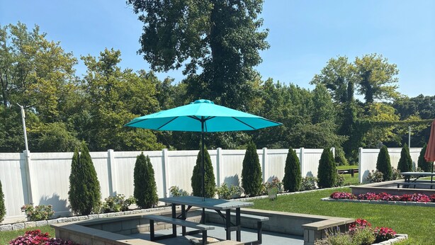A patio area with a picnic table and a turquoise umbrella, surrounded by flower beds and a white fence. - Red Roof Inn Wildwood - Cape May/ Rio Grande (Rio Grande)