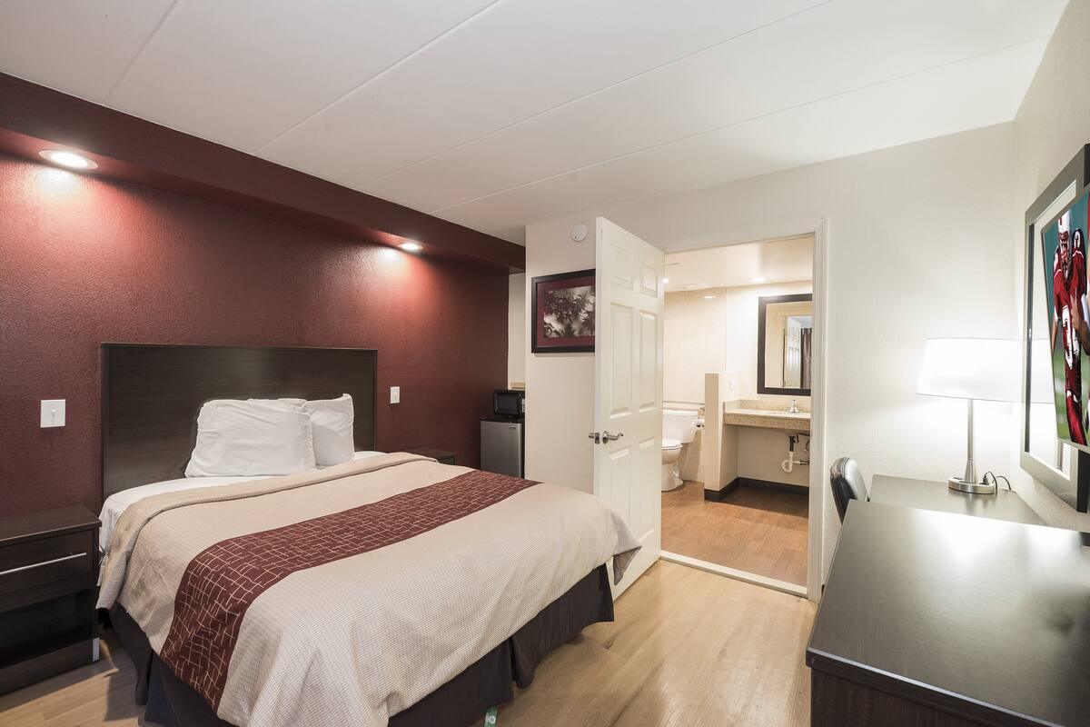A hotel room with a bed, bedside table, and a desk with a lamp.