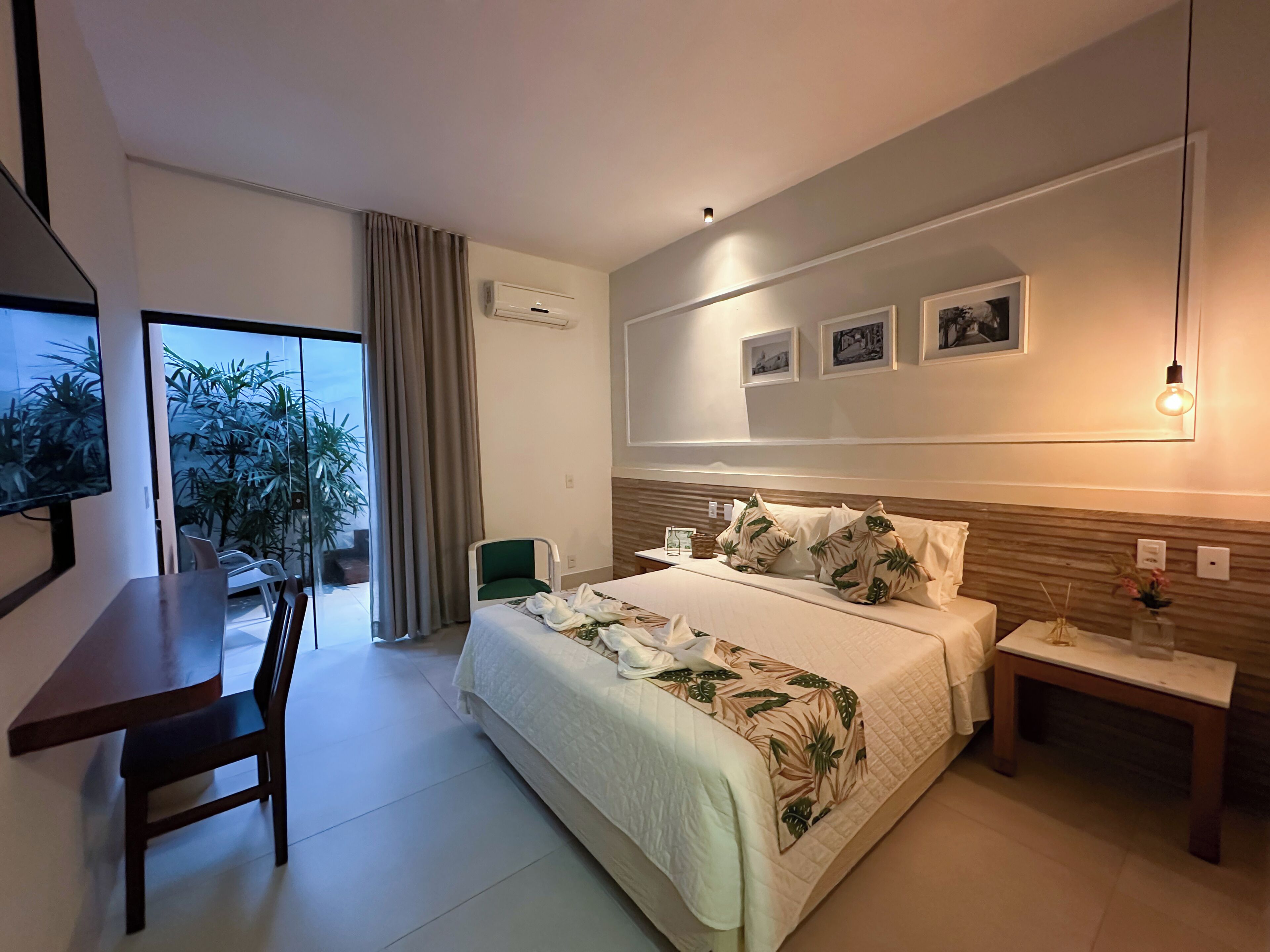 Ein modernes Hotelzimmer mit Bett, Schreibtisch, Stuhl und Blick auf Grün hinter einer Schiebetür.