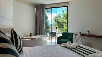 A hotel room with a bed, a small table, a chair, and a balcony with a view of trees and buildings.
