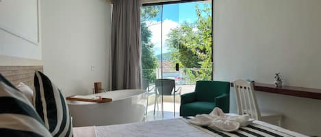 A hotel room with a bed, a small table, a chair, and a balcony with a view of trees and buildings.