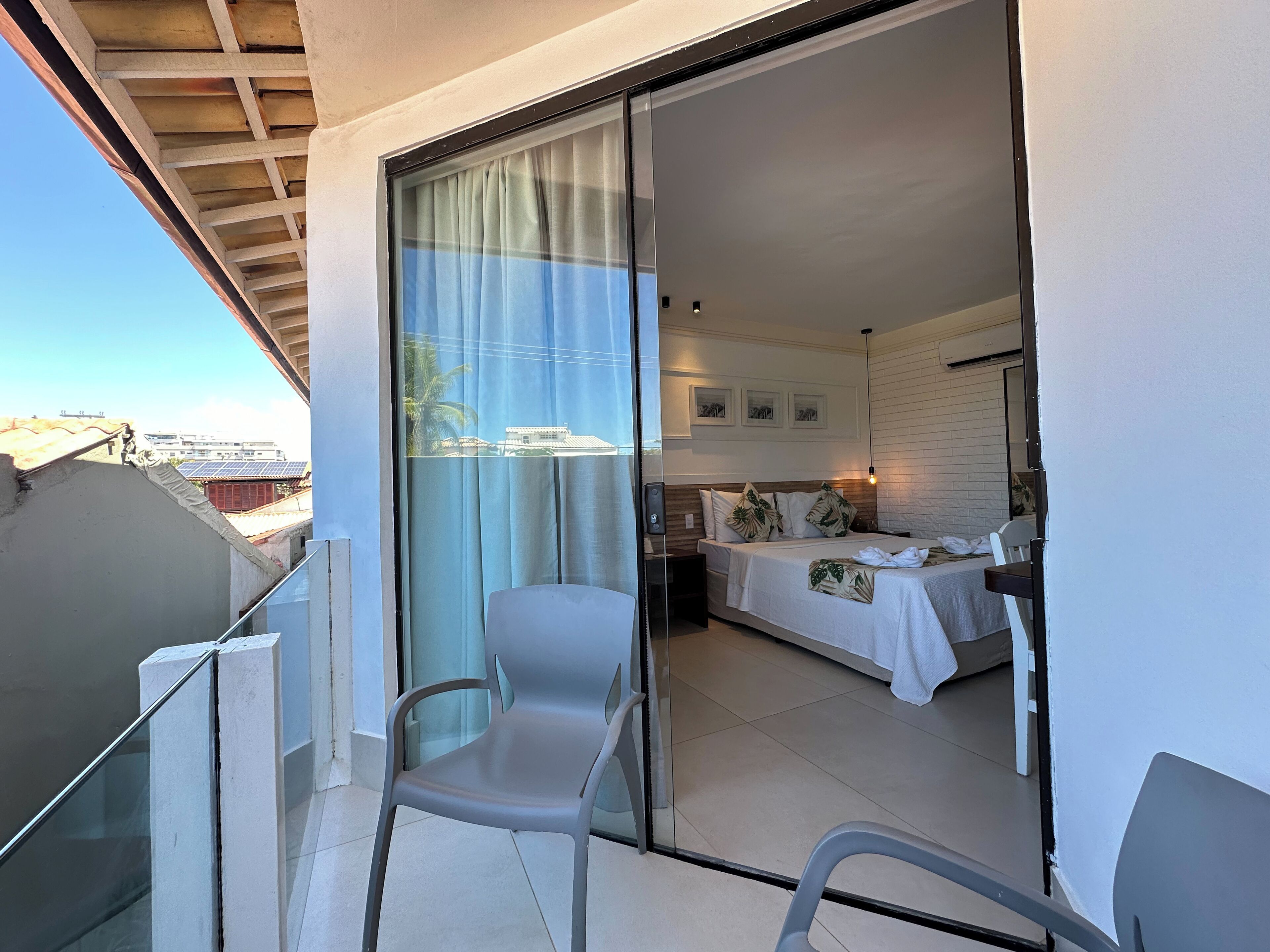 A balcony with a chair, a bed with a white bedspread and pillows, and a view of buildings through a glass door.