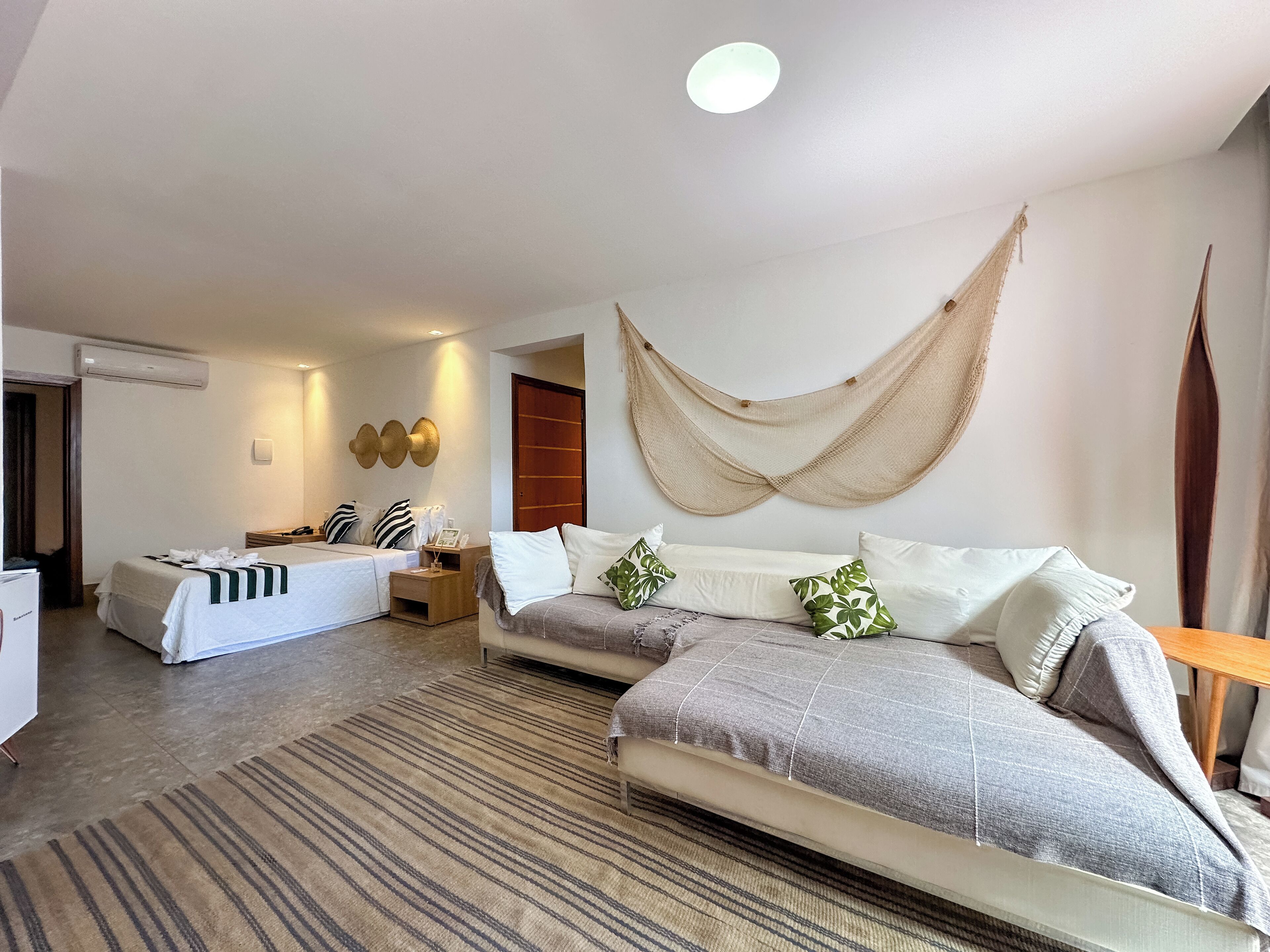 A hotel room with a large bed, a smaller bed, a sofa, a wooden side table, a wall-mounted air conditioner, and a hanging hammock.