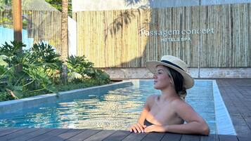 Una mujer descansando junto a una piscina con cubierta de madera, rodeada de plantas tropicales y una cerca de bambú.