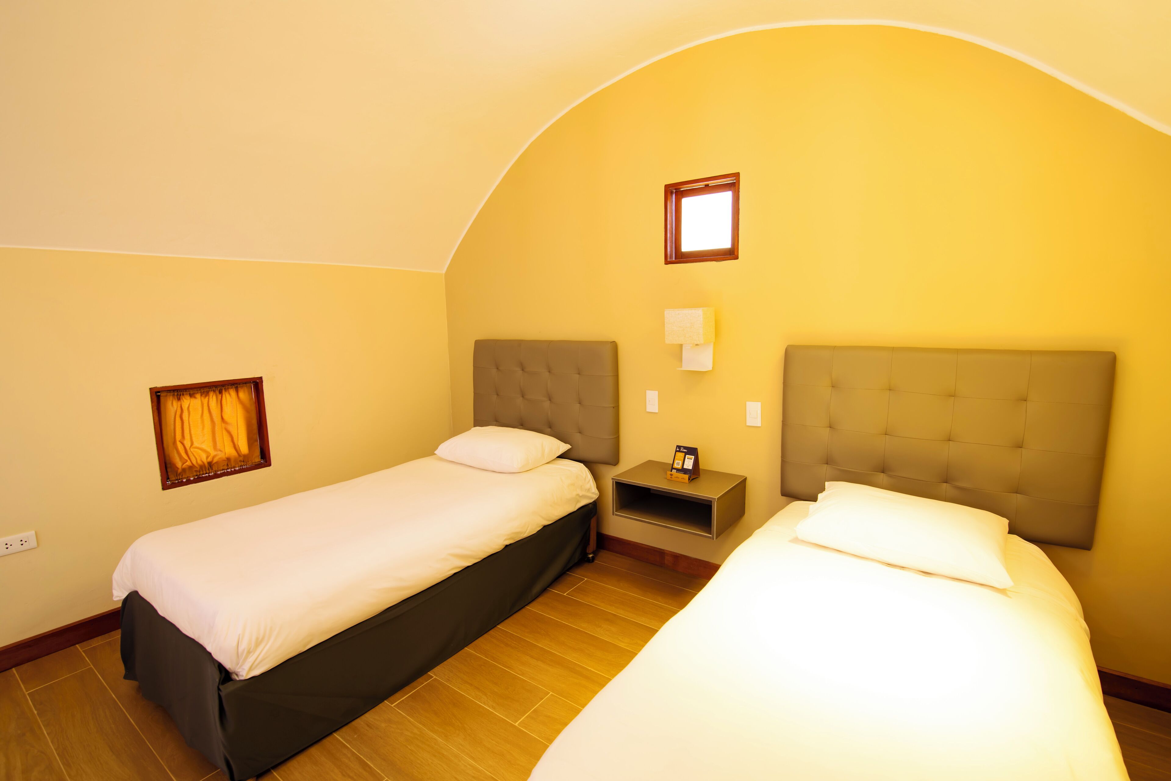 A hotel room with two beds, a small bedside table with a lamp, and a window with curtains.