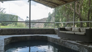 A hot tub area with a stone wall, a wicker sofa, and a view of a mountainous landscape through large windows. - El Refugio Hotel Colca (Chivay)