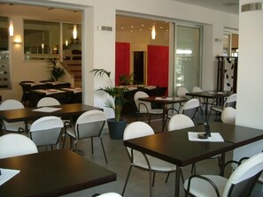 A modern dining area with multiple tables and chairs, a potted plant, and a red accent wall. - Hotel Emperador (Jesolo)