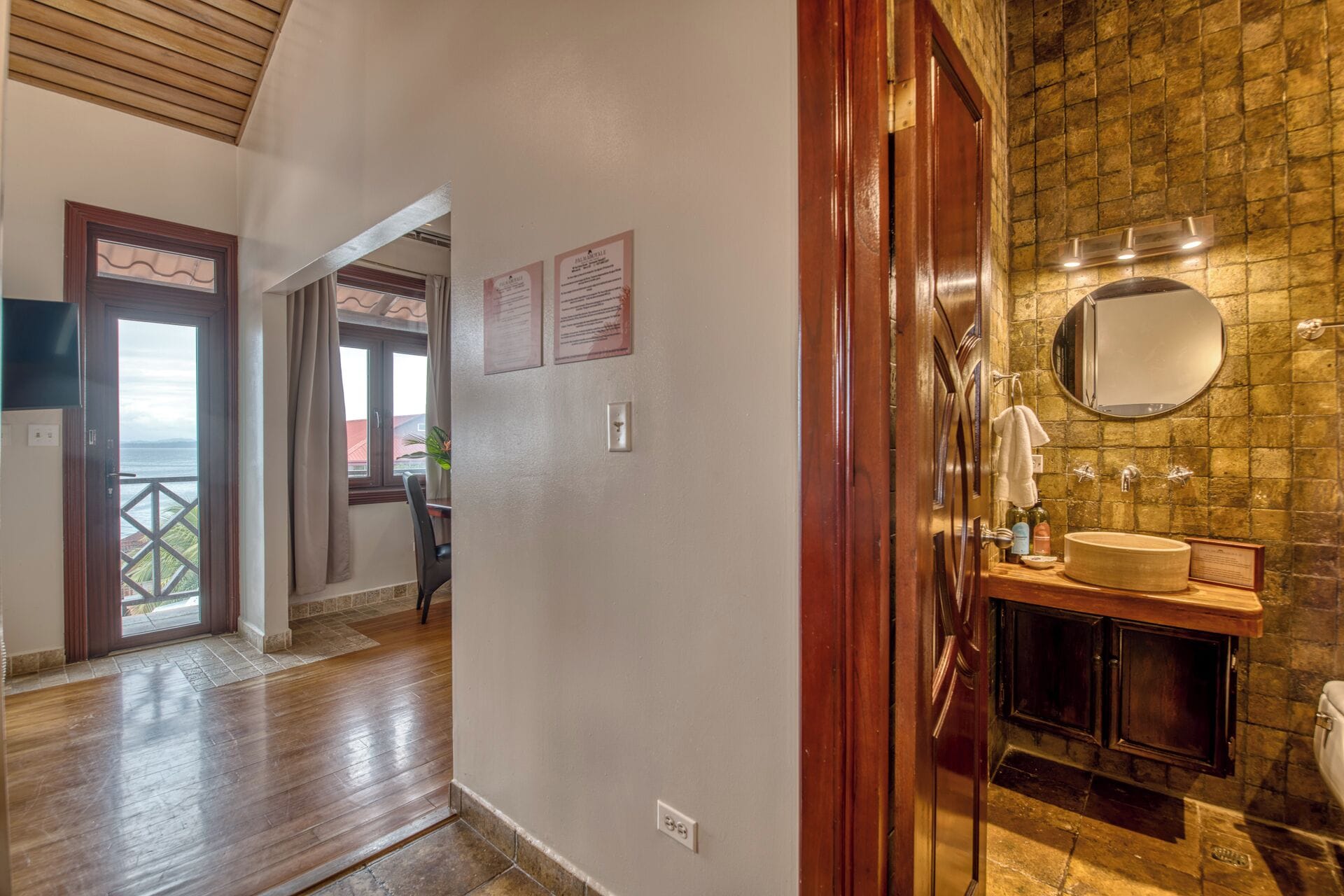 A hotel hallway leading to a bathroom with a stone wall, a wooden door, and a view through a window.