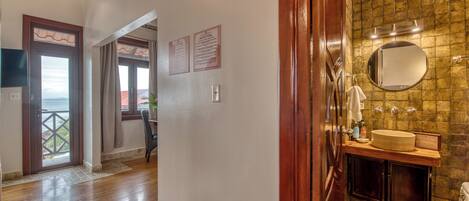 A hotel hallway leading to a bathroom with a stone wall, a wooden door, and a view through a window.