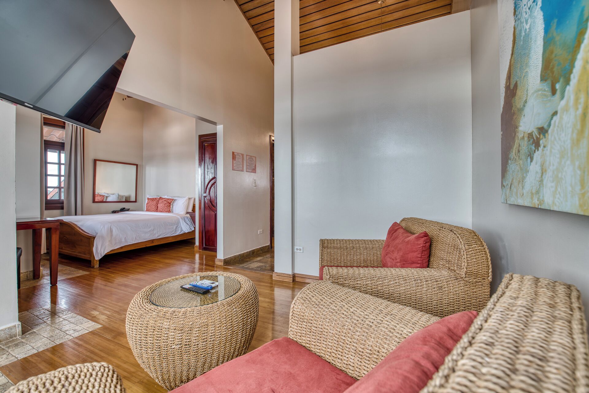 Une chambre d’hôtel moderne, avec un lit, un canapé en osier, un pouf rond et un grand tableau accroché au mur.