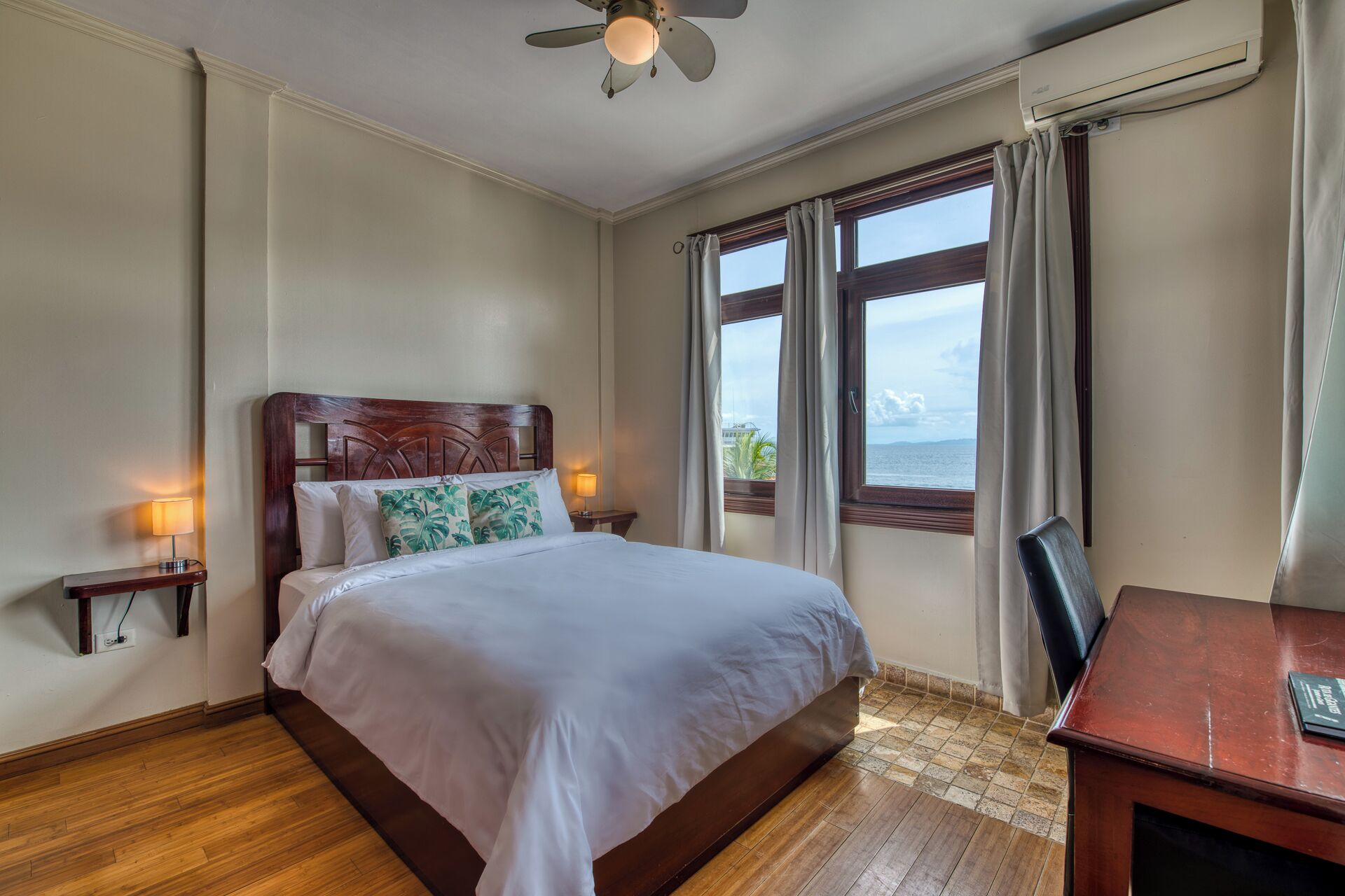 A hotel room with a bed, a desk, a chair, a nightstand, a lamp, a ceiling fan, and a view of the sea.
