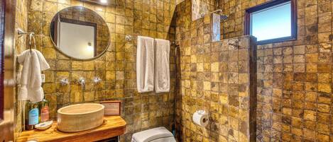 A bathroom with a stone wall, a round mirror, a sink with a wooden countertop, a toilet, and a shower with a window.