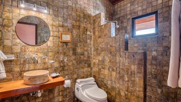 A bathroom with a stone wall, a round mirror, a sink, a toilet, and a shower.