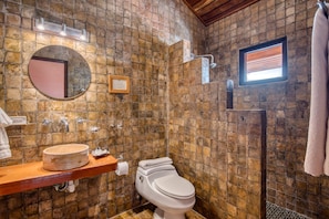A bathroom with a stone wall, a round mirror, a sink, a toilet, and a shower. - Hotel Palma Royale (Bocas del Toro)