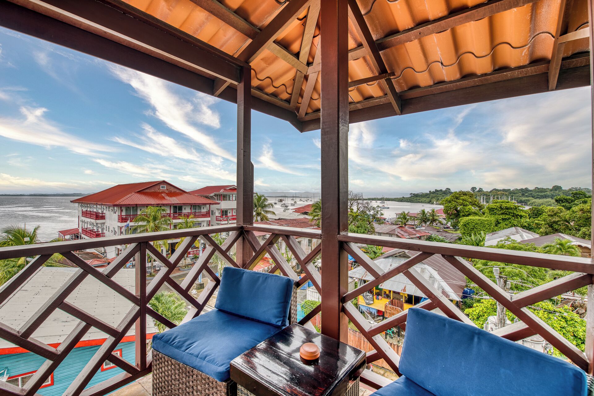 Un balcon avec deux chaises bleues dotées de coussins et une petite table, donnant sur une zone côtière avec des bâtiments et une vue sur la mer.