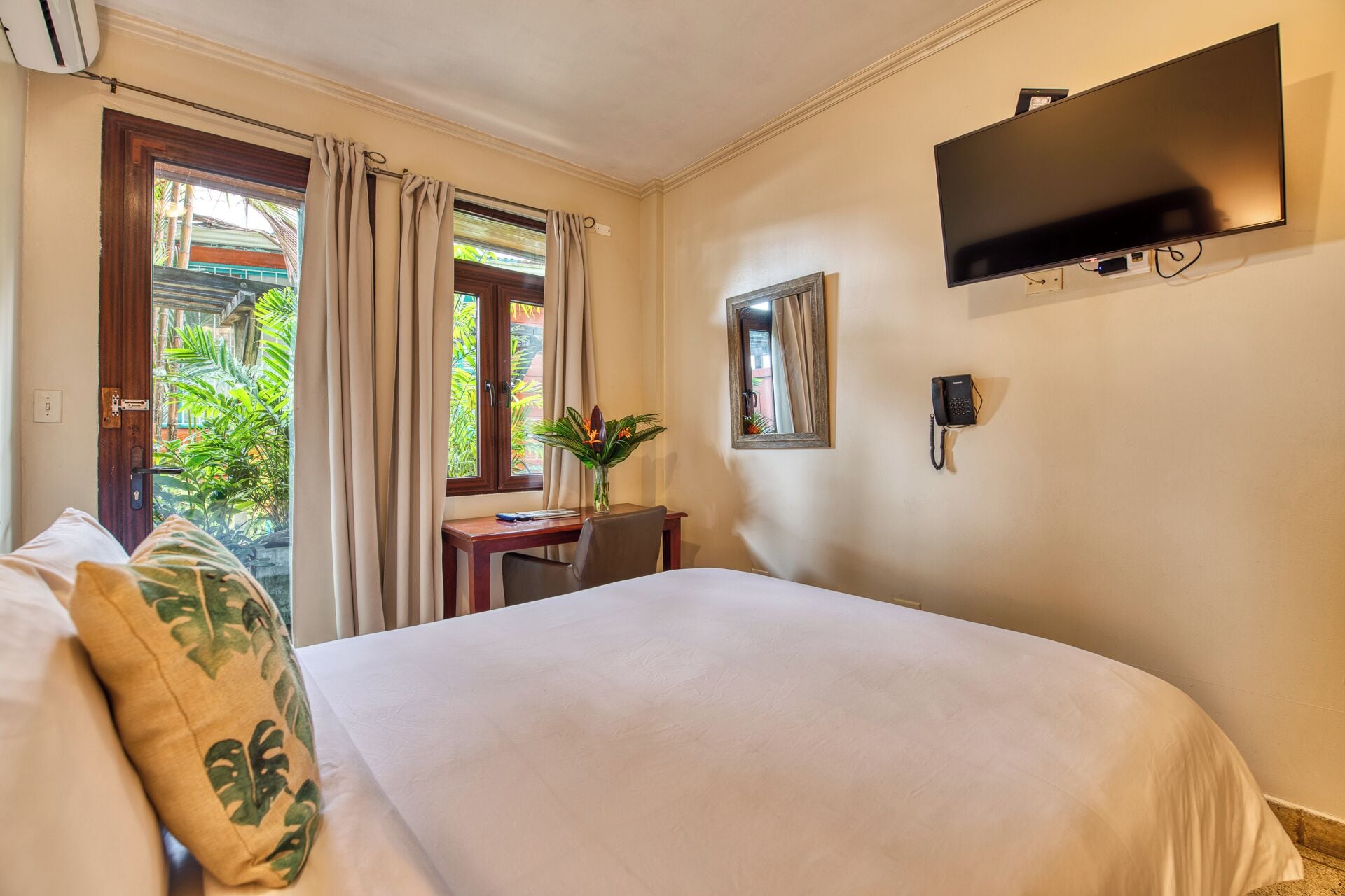 Une chambre d’hôtel comprenant un lit, un bureau avec une chaise, un téléphone, une télévision et une fenêtre avec des rideaux.